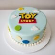 Classic pale-blue Toy Story cake with central logo, white cloud shapes, green and yellow top accents, colourful side ovals and white pearl border