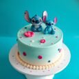 Pastel blue Stitch cake with two blue character figures, pink tropical flowers, colourful dots and a cream shell border