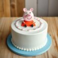 Peppa Pig cake with seated 3D character topper, smooth white icing, pink dotted border and blue cake board