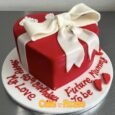 Red heart-shaped gift box cake with oversized white fondant bow, wrapped ribbon, small heart and baby-footprint decorations, and custom board message