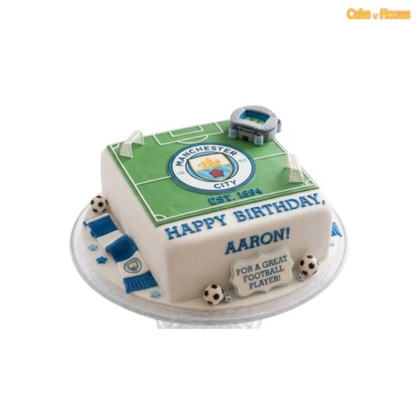 Square Manchester City football pitch cake with green field, club crest, miniature stadium, white goals, blue scarf and football decorations