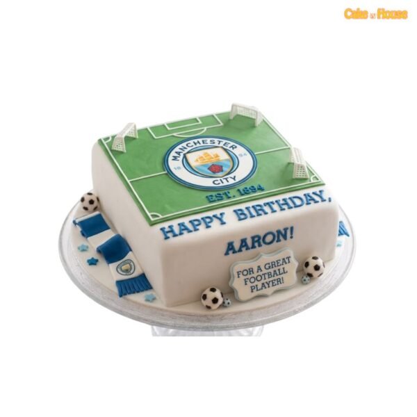 Square Manchester City football pitch cake with green field, white markings, club crest, three goal posts, blue scarf and miniature footballs