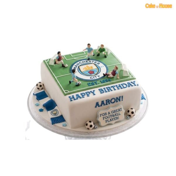 Square Manchester City football pitch cake with green field, central club crest, player figurines, white goals, blue scarf and football decorations